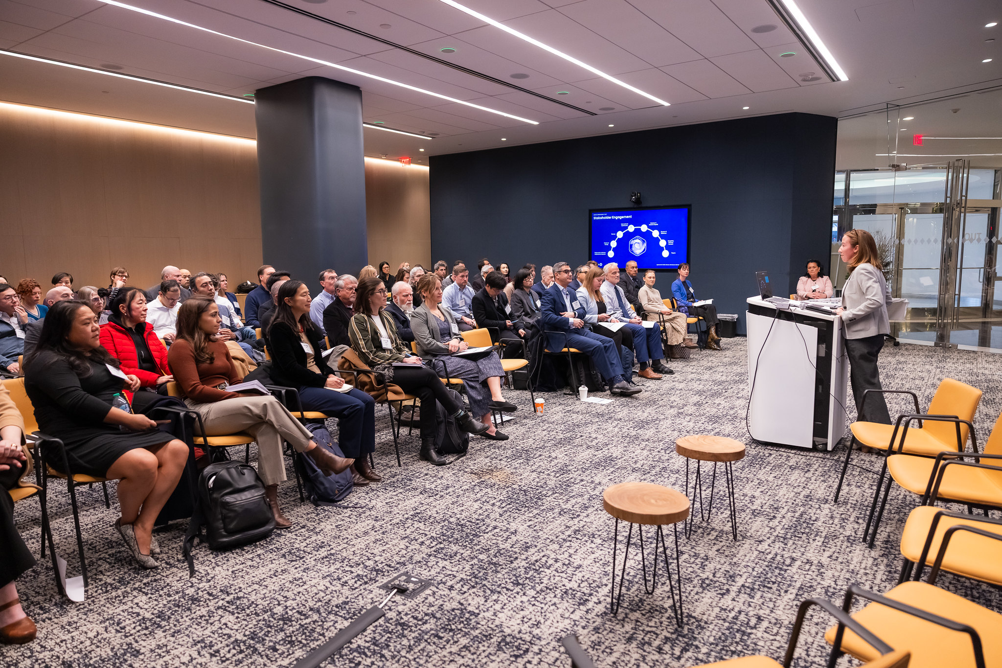 A room full of people listens to a speaker at a podium.